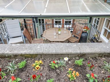 A small outdoor patio with a wooden table and chairs a bottle and glasses on the table and a flower bed in the foreground at Blacksmith Cottage in Appleby-in-Westmorland