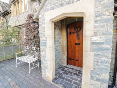 A wooden door with a heart decoration in a stone wall next to a white metal bench on a paved area at Bears Cottage in Bretforton near Broadway