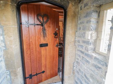 A wooden front door partially open with black metal hinges and a heart-shaped wreath at Bear's Cottage in Bretforton near Broadway