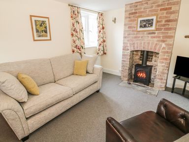 A living room with a beige sofa and cushions a brown armchair a brick fireplace with wood stove a window with floral curtains and framed pictures at Bears Cottage in Bretforton near Broadway