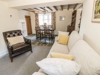 A living room with a beige sofa and brown armchair next to a wooden dining table with chairs and floral curtains at Bears Cottage in Bretforton near Broadway