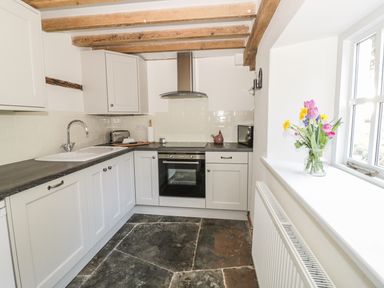 A kitchen with white cabinets a black countertop a sink an oven a range hood a toaster a microwave and flowers on the windowsill at Bears Cottage Bretforton near Broadway