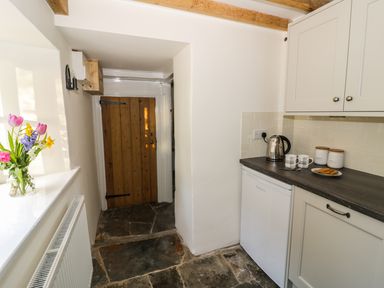 A small kitchen area with a wooden door stone floor a kettle mugs and jars on the countertop and flowers on a windowsill at Bears Cottage in Bretforton near Broadway