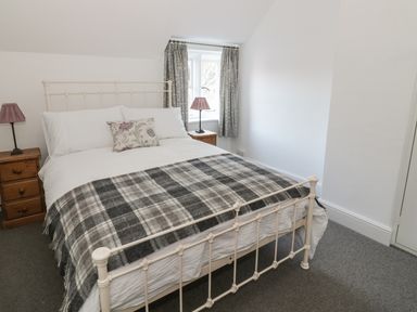 A bedroom with a metal bed frame gray and white plaid blanket two wooden bedside tables with lamps and a window with curtains at Bears Cottage in Bretforton near Broadway