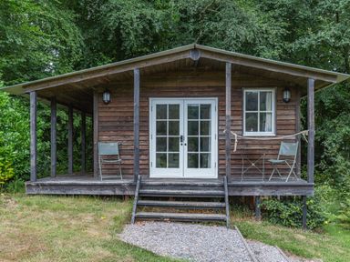 A wooden cabin with a porch two chairs a small table and steps leading to a gravel path at Lakeside Cabin in Sheldon Blackdown Hills
