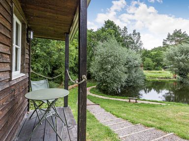 A wooden porch with a green table and chair overlooking a pond and trees at Lakeside Cabin in Sheldon Blackdown Hills