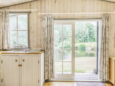A kitchen area with a sink and dish rack next to a glass door overlooking a pond and greenery at Lakeside Cabin in Sheldon Blackdown Hills