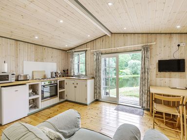 A kitchen and living area with wooden floors walls and ceiling with a sofa dining table tv and door opening to a view of a lake at Lakeside Cabin in Sheldon Blackdown Hills