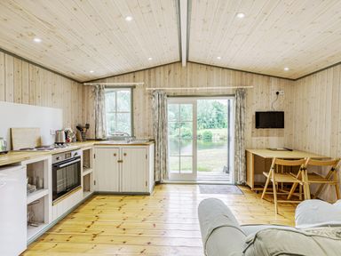 A kitchen and dining area with wooden walls and floor and a view of a lake through glass doors at Lakeside Cabin in Sheldon Blackdown Hills