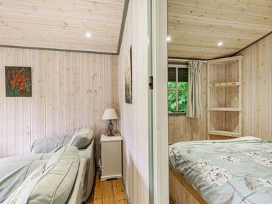 A room with a light green couch and small dresser with lamp next to a bedroom with bed and corner shelves at Lakeside Cabin in Sheldon Blackdown Hills