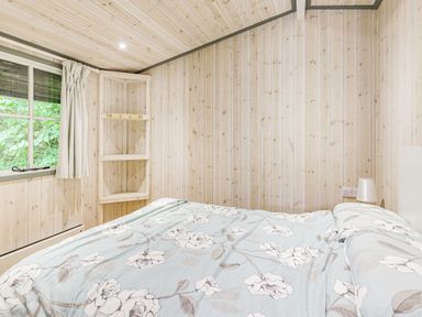 A bedroom with wooden walls a window with curtain a corner shelf a bed with floral bedding and a bedside lamp at Lakeside Cabin in Sheldon Blackdown Hills