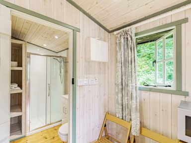 A bathroom with a glass shower enclosure and wooden shelving next to a small room with a wooden chair and an open window at Lakeside Cabin in Sheldon Blackdown Hills
