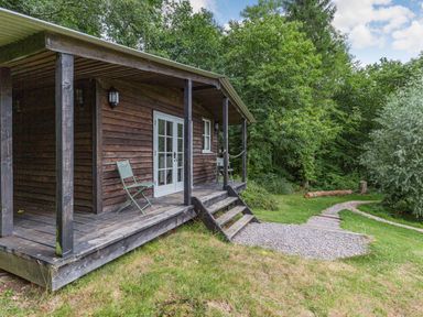A wooden cabin with a covered porch and two green chairs beside a gravel path in a wooded area at Lakeside Cabin in Sheldon Blackdown Hills