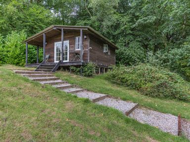 A small wooden cabin on stilts with a covered porch and steps leading up to it surrounded by trees and grass at Lakeside Cabin in Sheldon Blackdown Hills