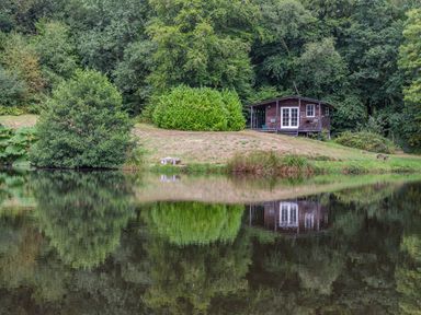 A wooden cabin on a grassy slope near a pond with trees and bushes in Sheldon Blackdown Hills