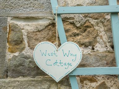 A heart shaped sign reading West Wing Cottage hanging on a blue wooden frame against a stone wall at West Wing Cottage in Hexham