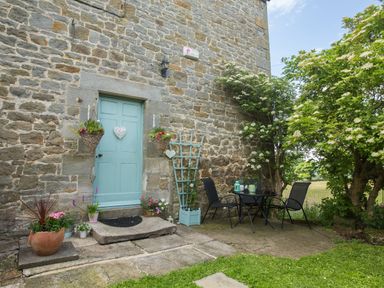 An outdoor patio area with a blue door on a stone wall two black chairs and a table with plants and flowers at West Wing Cottage in Hexham
