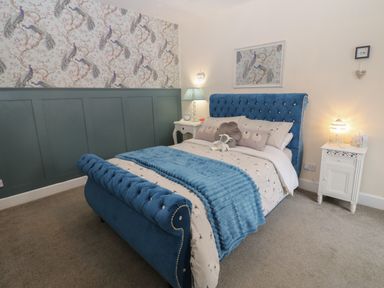 A bedroom with a blue upholstered bed white bedside tables and peacock wallpaper at West Wing Cottage in Hexham