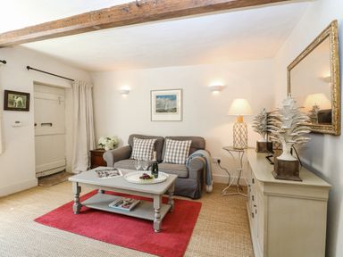 A living room with a gray sofa two plaid pillows a coffee table with a tray and glasses a red rug a cabinet with decorative items and a lamp at Bridge Cottage in Aylsham