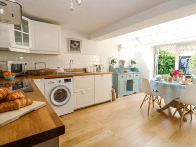 A kitchen with wooden countertops white cabinets a washing machine a blue sideboard and a dining table with chairs at Bridge Cottage in Aylsham