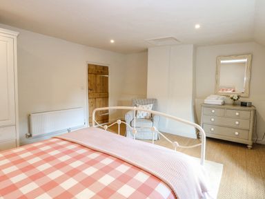 A bedroom with a bed covered in pink and white checkered bedding a wooden door a checkered armchair and a dresser with a mirror at Bridge Cottage in Aylsham