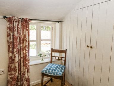 A wooden chair with a blue cushion next to a window with patterned curtains and white paneled closet doors at Bridge Cottage in Aylsham