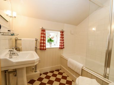 A bathroom with a pedestal sink towel racks bathtub with glass shower screen and a window with red check curtains and flowers at Bridge Cottage in Aylsham