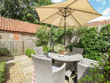 An outdoor patio with a glass table, four woven chairs, a large umbrella, and greenery around at Bridge Cottage in Aylsham