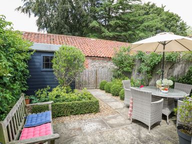 A garden patio with a wooden bench with cushions a round glass table with wicker chairs and an umbrella at Bridge Cottage in Aylsham
