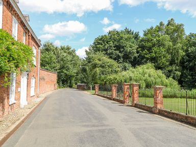 A road lined with a brick house and a brick fence with metal railings next to green trees and grass at Bridge Cottage in Aylsham