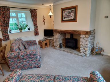 A sitting room with patterned sofas a stone fireplace a window with curtains and a television at Magpie Cottage South Brewham