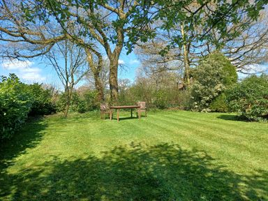 An outdoor garden area with two wooden chairs and a wooden table on grass surrounded by trees and shrubs at Magpie Cottage South Brewham