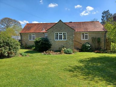 A stone cottage with red roof tiles and white framed windows surrounded by green bushes and lawn at Magpie Cottage in South Brewham
