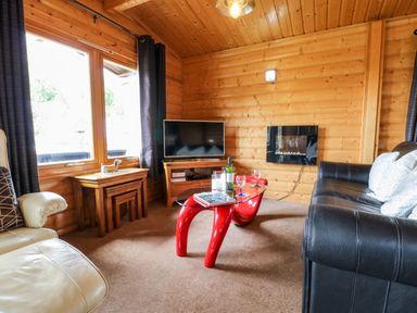 A living room with wooden walls and ceiling a black leather sofa a cream armchair a red glass coffee table a television and nested wooden tables at Heathcliff in Northallerton