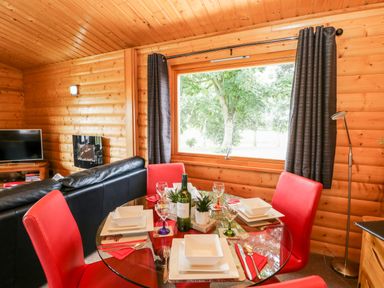 A dining area with red chairs and a glass table set in a wooden cabin with a large window showing trees outside at Heathcliff in Northallerton