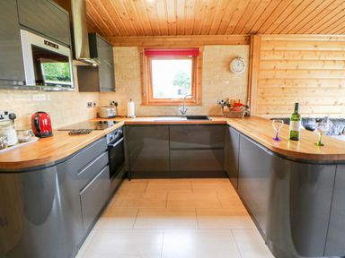 A kitchen with grey cabinets wooden countertops a window with a red blind and a bottle of wine on the counter at Heathcliff in Northallerton