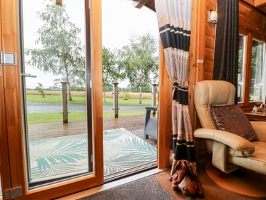 A sliding glass door opening to a wooden deck with a mat and trees outside by a leather chair with a cushion and curtain at Heathcliff in Northallerton