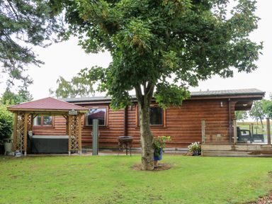A lawn with a tree a wooden cabin a gazebo and a deck with glass railing at Heathcliff in Northallerton