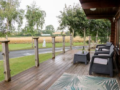 A wooden deck with three chairs and a table with a wine bottle and glasses overlooking a grassy yard and trees at Heathcliff in Northallerton