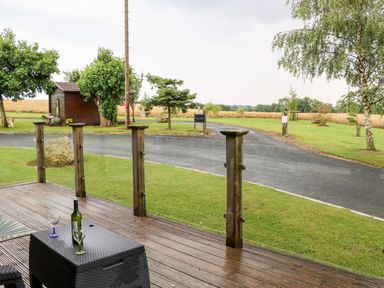 A wooden deck with a table holding a bottle and two glasses overlooking a paved driveway and grassy area with trees and a small wooden shed at Heathcliff in Northallerton