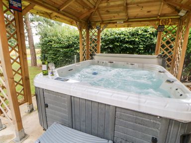 An outdoor hot tub with bubbling water under a wooden gazebo with lattice sides and a bottle of wine with glasses at Heathcliff in Northallerton