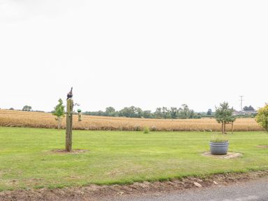 A grassy field with a wooden post holding a bird feeder and a bird decoration next to a flower pot and a field of crops at Heathcliff in Northallerton
