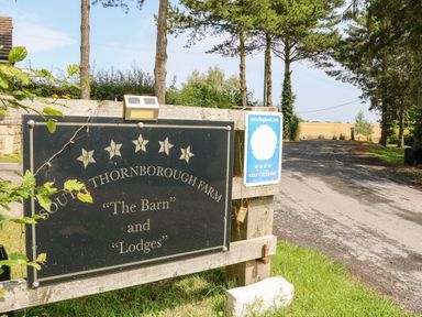 A roadside sign for South Thornborough Farm The Barn and Lodges with trees and a rural road in the background