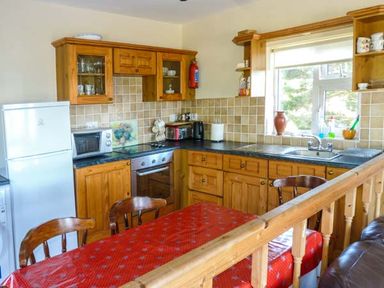 A kitchen with wooden cabinets a white refrigerator a microwave stove and a red tablecloth on the dining table at Ti Sheamuis in Cashel near Carna County Galway