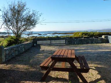 A wooden picnic table in a gravel yard with a stone wall and gate overlooking the sea at Ti Sheamuis Cashel near Carna County Galway