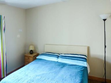 A bedroom with a bed with blue bedspread and striped pillows a wooden nightstand with a lamp and a floor lamp near a wall with colorful curtains at Ti Sheamuis Cashel near Carna County Galway