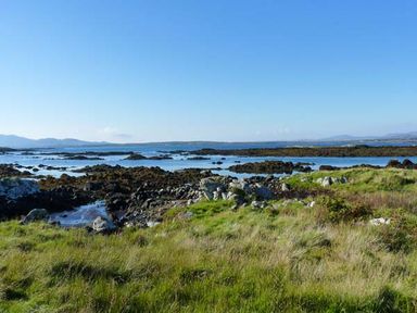 A rocky shoreline with grass in the foreground and water with distant hills in the background at Ti Sheamuis in Cashel near Carna County Galway