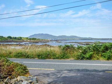 A coastal road with grass and bushes alongside and mountains visible across the water at Ti Sheamuis Cashel near Carna County Galway