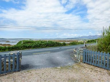 A road with a blue wooden gate and stone walls overlooking water and distant mountains at Ti Sheamuis Cashel near Carna County Galway