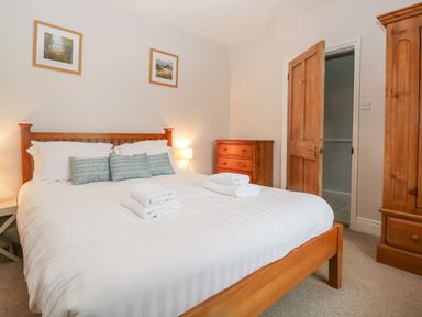 A bedroom with a wooden bed frame white bedding folded towels wooden dresser wardrobe and bedside lamp at Fishers Retreat in Bowness-On-Windermere